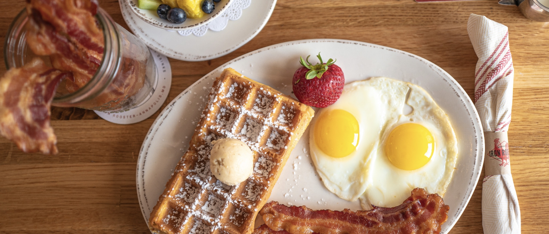 Full table breakfast at Iron Rooster including coffee, a bowl of fresh fruit, jar of Iron Rooster's famous candied bacon and a plate with Belgian waffles and two sunny side up eggs.